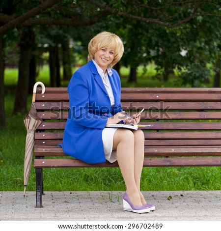 Mature business blonde woman in a jacket with diary sitting on a bench in a summer park