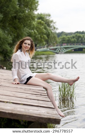 Young beautiful blonde woman in a white tunic sits on a wooden pier on the lake