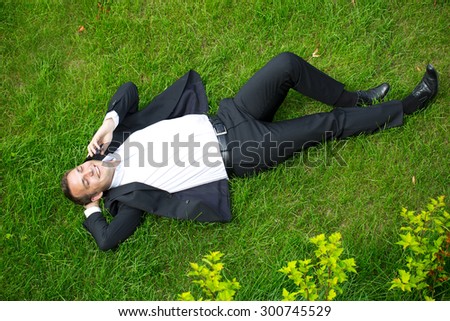 Cheerful young businessman calling by phone lying on green grass