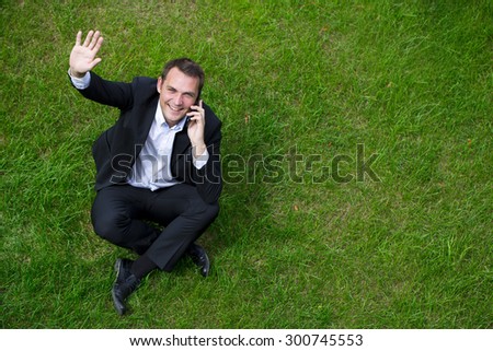 Cheerful young businessman calling by phone sitting on green grass