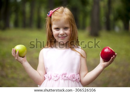 Happy little red-haired girl in a pink dress holding two apples, summer green park