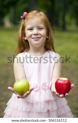 Happy little red-haired girl in a pink dress holding two apples, summer green park
