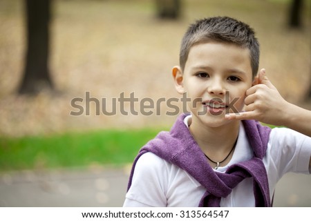 Little Boy making a call me gesture, against background of summer park