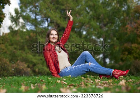 Young beautiful woman in red leather jacket and blue jeans sitting in the autumn park