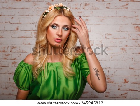 portrait of a glamorous girl in a green dress on brick wall background.girl in floral wreath and with a tattoo on his arm. glamorous blonde with a tattoo in a green dress on brick wall background