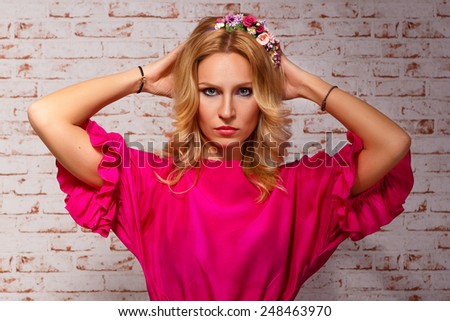 Glamour girl in a pink dress holding her hands behind her head. blonde in floral wreaths brick wall background. Cut Model Woman. Perfect Skin. Professional Make-up.