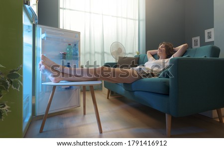 Happy woman lying on the sofa at home and cooling herself in front of the open fridge