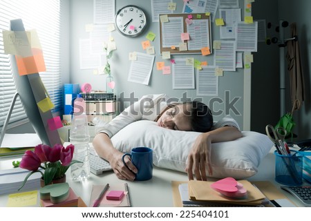 Tired businesswoman at office desk waking up with pillow and coffee.