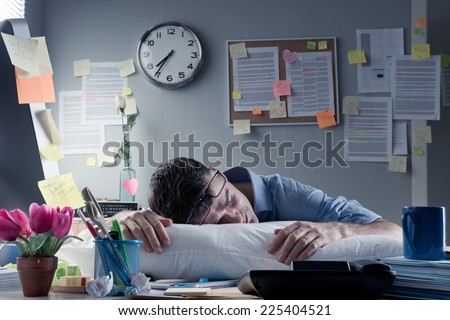 Exhausted businessman sleeping at workplace with a pillow on his desk.