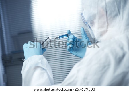 Researcher in safety hazmat suit preparing a syringe for injection with glass test tube.