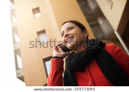 Smiling woman in red coat having a phone call with her smart phone, city buildings on background
