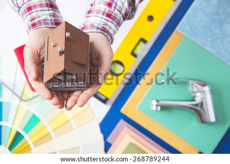 Home insurance, repair and decorating service concept with cupped hands holding a model house and faucet, color swatches and work tools on background