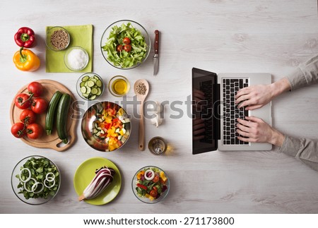 Man in the kitchen searching for recipes on his laptop with food ingredients and fresh vegetables on the left, top view