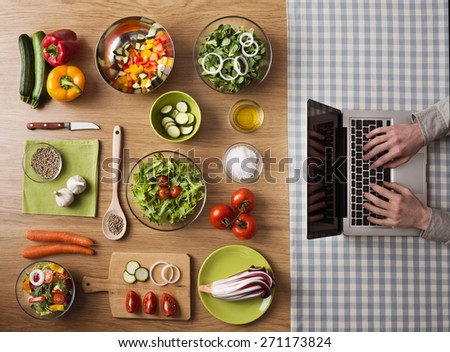 Vegetarian healthy food preparation at home on kitchen table with hands typing on a laptop on the right, top view