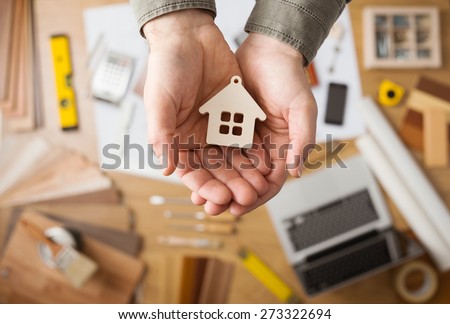 Real estate agent holding a small house, desktop with tools, wood swatches and computer on background, top view