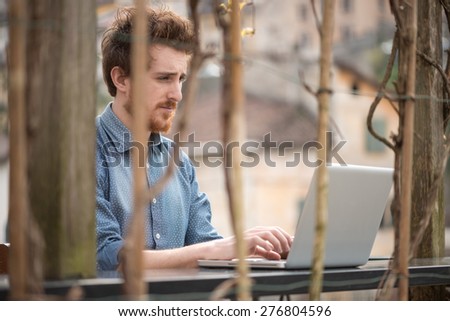 Young hipster man working on a laptop on an outdoor table with city on background