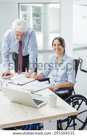 Confident business woman in wheelchair working at office desk with her male colleague and smiling at camera