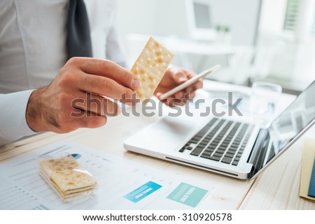 Businessman having a snack at desk holding a cracker and a smart phone, hands close up, unrecognizable person