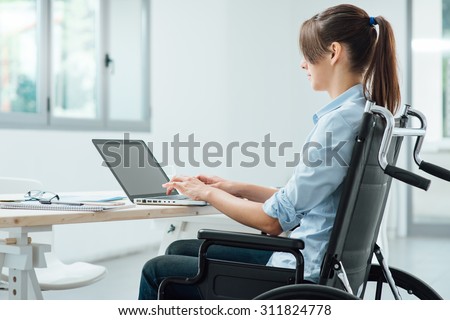 Young disabled business woman in wheelchair working at office desk and typing on a laptop, accessibility and independence concept