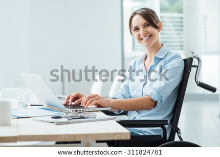 Confident happy businesswoman in wheelchair working at office desk and using a laptop, she is smiling at camera, disability overcoming concept