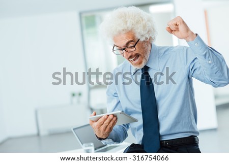 Cheerful successful business man with raised fist receiving good news on a digital tablet, he is leaning on office desk, room interior on background