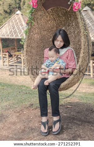 young mother holding baby on rattan swing. Baby play guitar toy in the mother hug