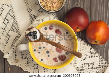 Muesli in a yellow ceramic cup with milk and berries on a dark wooden table with lace cotton napkin, peaches and wooden spoon
