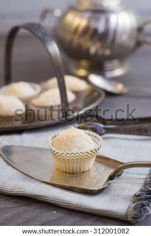Coconut protein cupcake sweets in a paper pack on a silver plate with silver spoons and tongs on a grey napkin on a dark grey wooden background