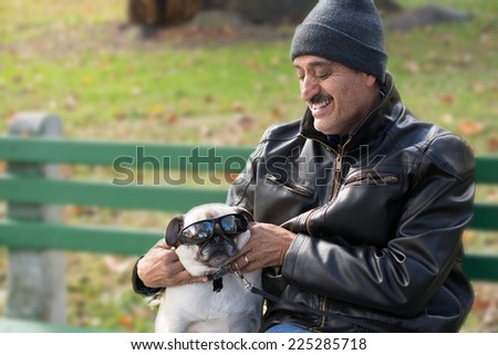 A Hispanic man sitting on a bench with a pug dog, enjoying playing with the dog and putting sunglasses on it.