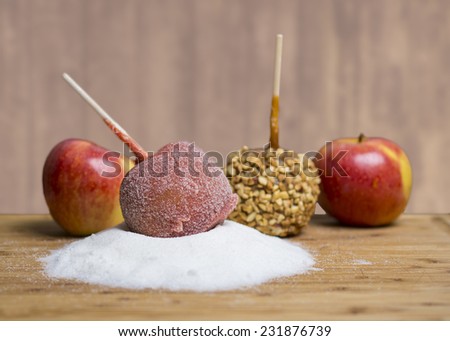 A candy apple coated sugar sits atop a pile of sugar. Another candy apple with chopped nuts behind it, with two Gala apples. On a wooden cutting board with a textured background.