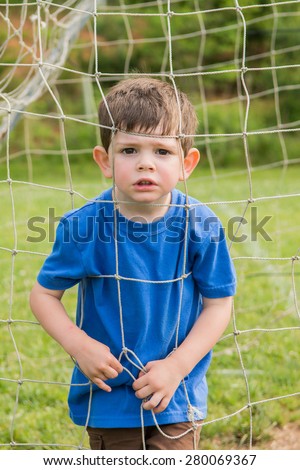 A little boy pressing his face into a goal net, outside on a spring day. He is making a serious face. Vertical.