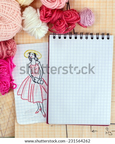 Fashion designer's opened notebook with a draft scetch fashion dress on the background of graph paper with balls of wool.