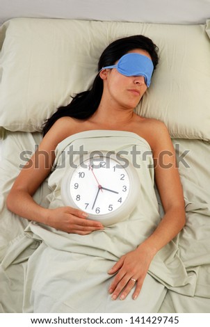 Beautiful sleeping young woman in sleep eye mask holding a clock.