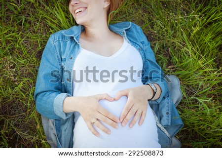 Smiling happy pregnant woman lying on the grass and makes a heart-shaped hands on her belly