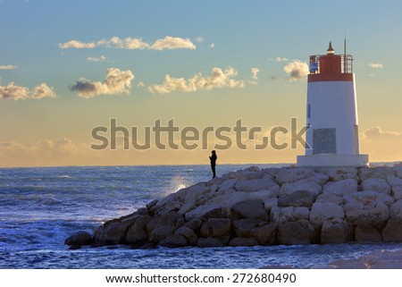 Lonely person in front of the sea on a sea wall near a small red and white lighthouse.