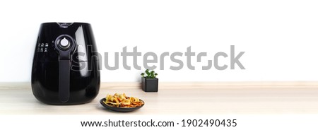 a black air fryer or oil free fryer appliance is on the wooden table in the kitchen with deep fried banan chips and small plant in the pot on white cement wall background during dinner in the party