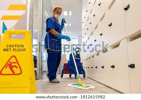 A cleaner with a mask on her face cleans the floor with the mop.Caution wet floor sign close up