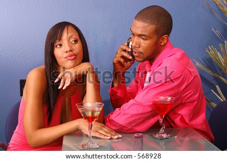 African American couple in a martini bar.Man talking on cell phone while the woman sits by annoyed and bored
