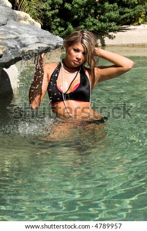 Sexy Caucasian woman wearing a black and pink bikini and wading in a swimming pool near a small waterfall