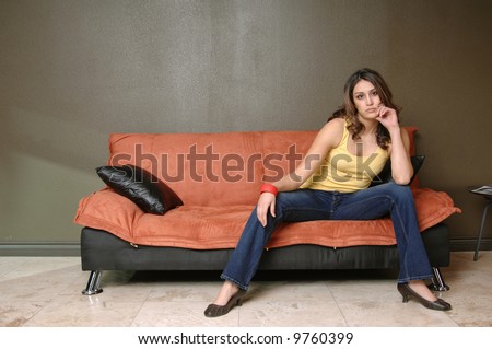 Pretty young Mexican woman in jeans and a yellow wife beater casually sitting on a black and orange couch with her chin in her hand