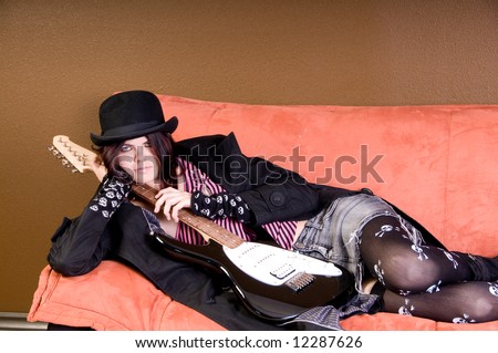 Beautiful and hip scene girl with red and black hair in a pink striped off the shoulder top, pirate sleeves and a felt bowlerwith an electric guitar relaxing on a couch