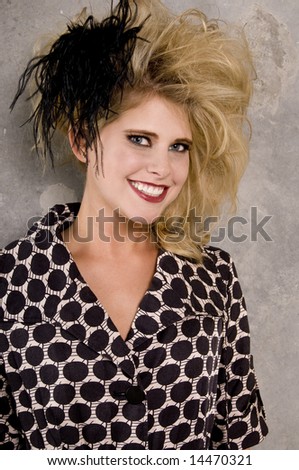 Smiling blonde woman wearing a high fashion mini dress with big teased hair,  and a feather hair piece against a concrete wall