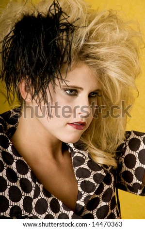 Young blonde woman wearing a high fashion mini dress with big teased hair, a small clutch and a feather against a yellow wall