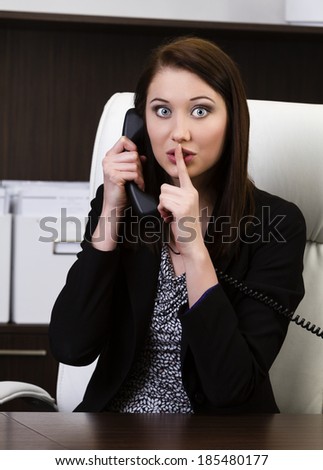 Portrait of attractive young business woman gesturing keep silence sign while talking on phone in the office