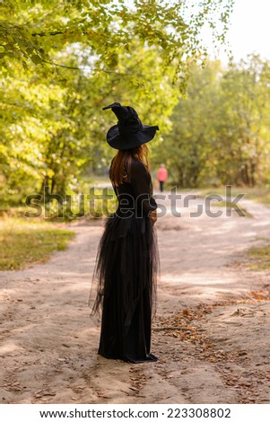 young long haired woman wearing black dress, black corset and witch\'s hat, standing back to the camera and looking away
