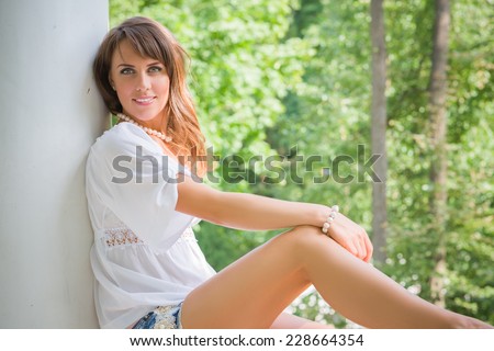Smiling long haired young woman in cutoffs and white tunic, sitting and leaning against a white pillar and looking at camera, trees in blurry background