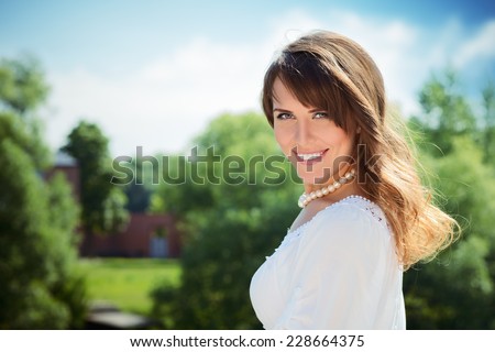 Smiling long haired young woman in white tunic and pearls necklace, looking at camera, trees and mansion in background