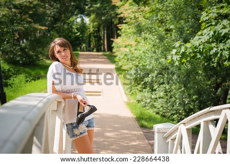 Smiling long haired young woman in white tunic and cutoffs, leaning against a white plank bridge railing and looking at camera, stilettos in hand, trees and paved road in background