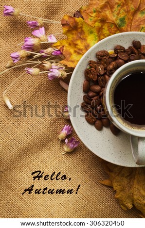 Tag with words hello autumn and the cup of coffee lying on the book with dry yellow maple leaves and everlasting flowers on sackcloth background