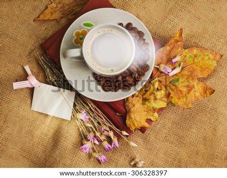 The cup of coffee lying on the books with dry yellow maple leaves and everlasting flowers on sackcloth background